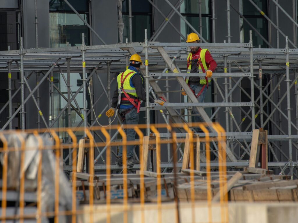 Two construction workers wearing safety gear and helmets working on scaffolding outdoors.