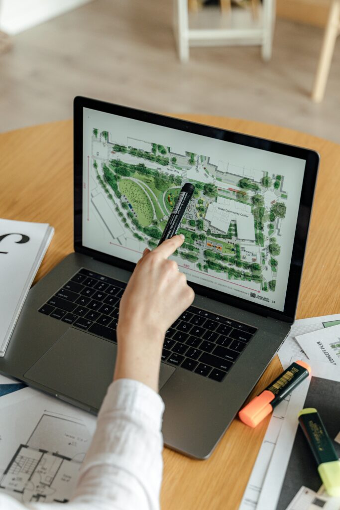 An architect reviews landscape design plans on a laptop, using highlighters and documents at a wooden desk.