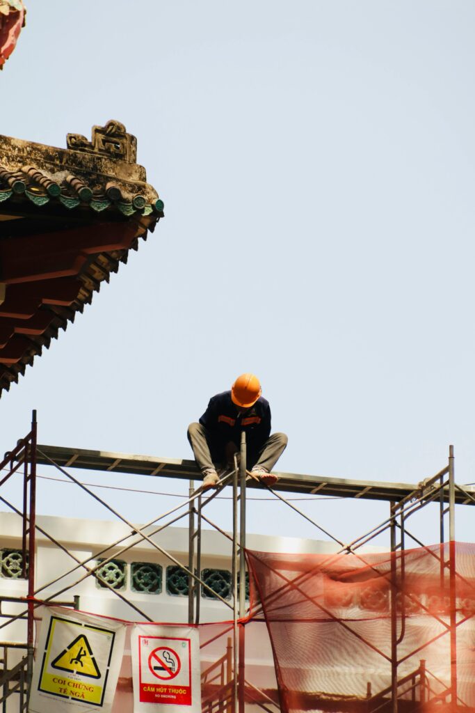 A construction worker wearing a helmet sitting on scaffolding under clear skies.